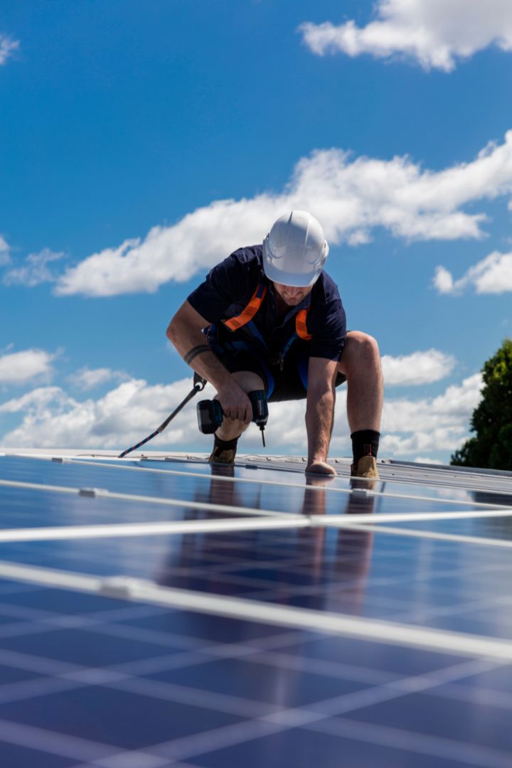 Trabajador con casco instalando paneles solares en el techo bajo un cielo azul con algunas nubes.